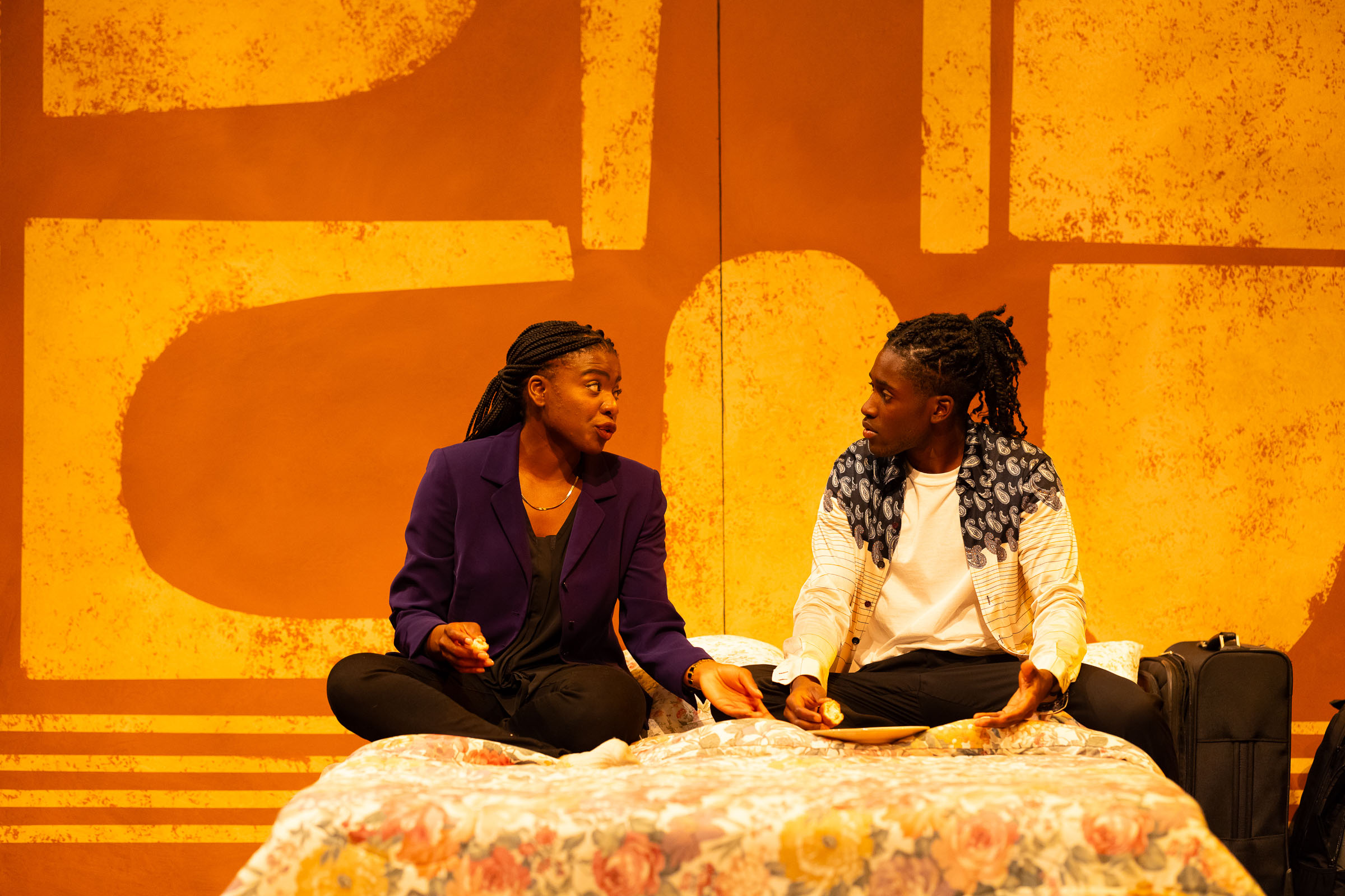 Two actors sit on a bed against an orange and yellow background